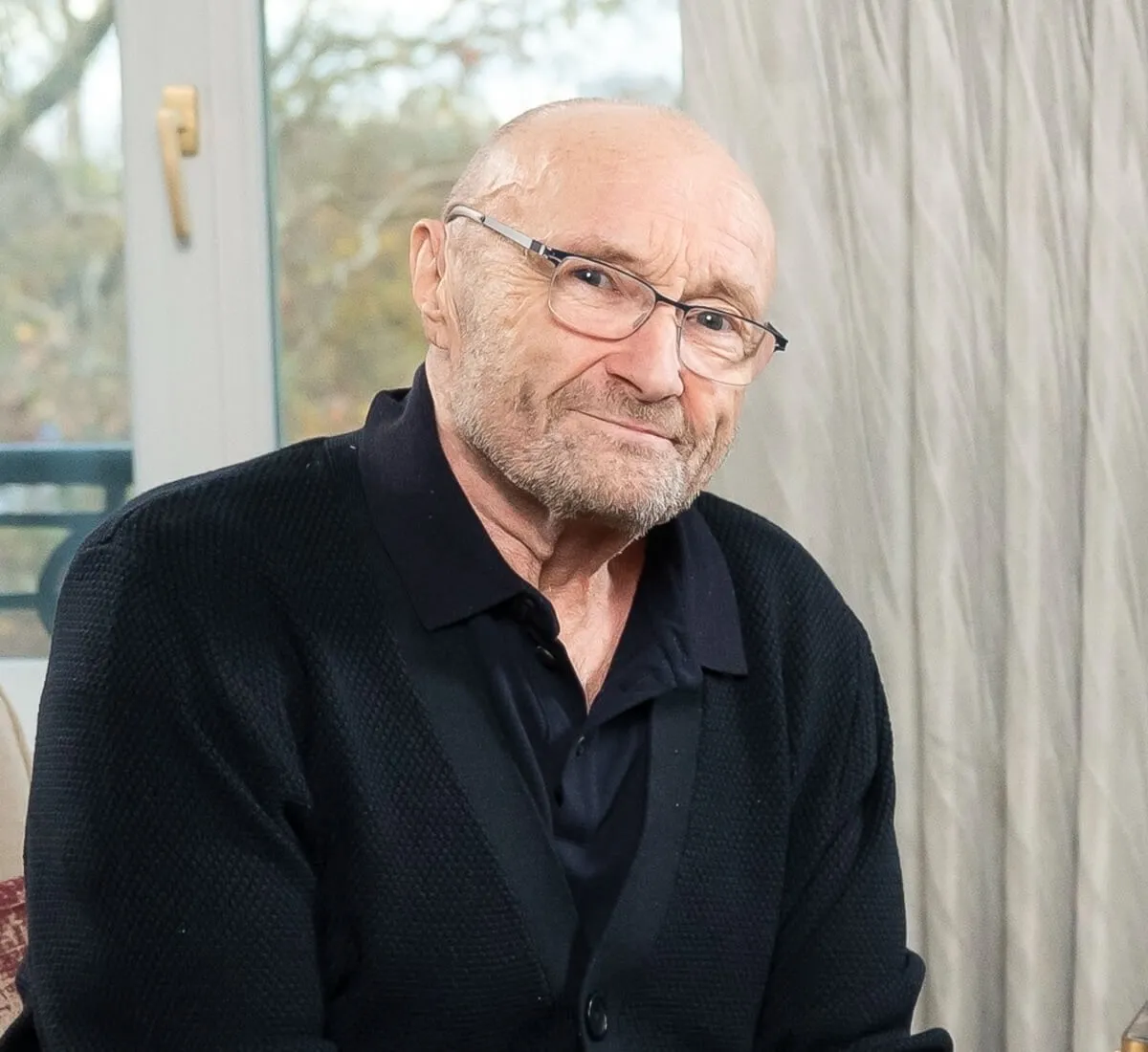 Phil Collins wears a black shirt and sits in front of a window.