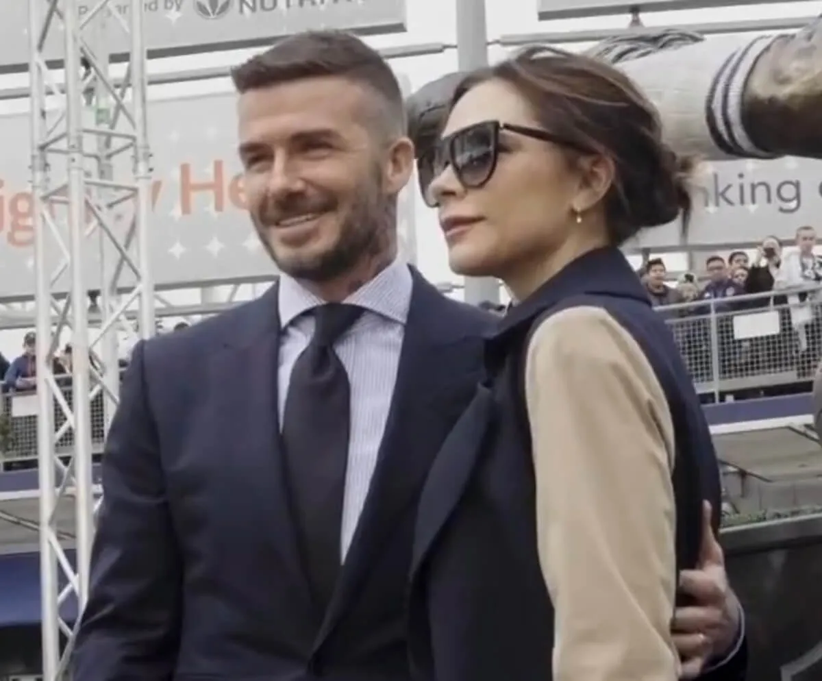 David Beckham wears a suit and stands with Victoria Beckham, who wears a coat and sunglasses.